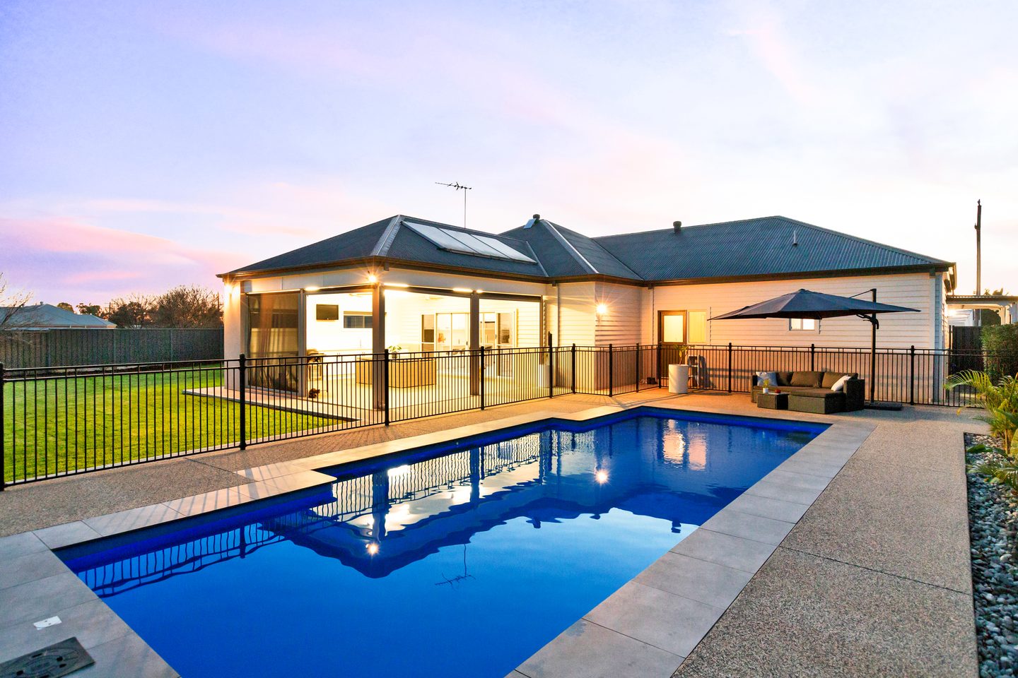 The heated in-ground pool beside the illuminated alfresco and rear lawn at dusk
